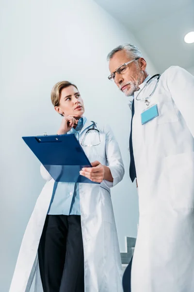 Vista de ángulo bajo del médico femenino que muestra el portapapeles al colega masculino en la escalera en el hospital - foto de stock