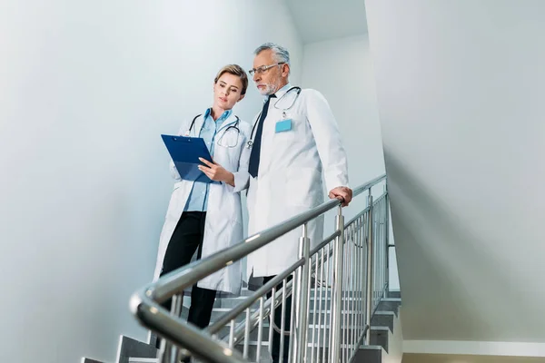 Female doctor showing clipboard to male colleague on staircase in hospital — Stock Photo