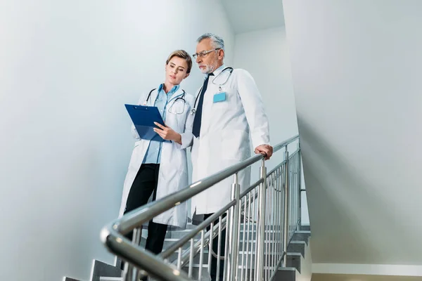 Doctora mostrando portapapeles a colega masculino en escalera en el hospital - foto de stock