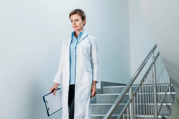 Doctora sonriente con portapapeles caminando por la escalera en el hospital - foto de stock