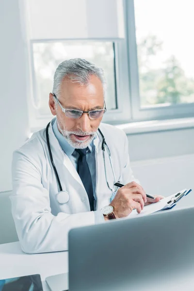 Arzt mittleren Alters schreibt in Klemmbrett und schaut im Büro auf Laptop — Stockfoto