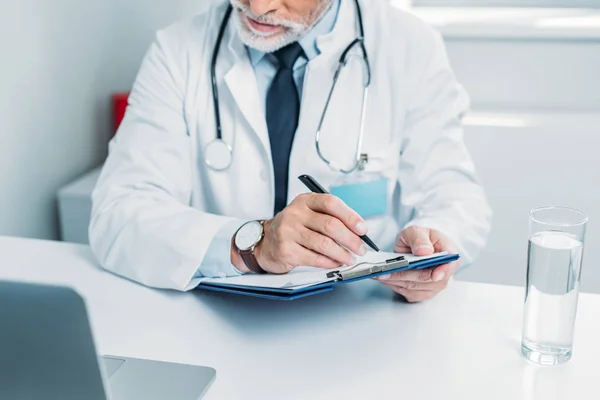 Partial view of male doctor writing in clipboard and looking at laptop in office — Stock Photo
