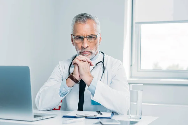 Médico masculino de mediana edad serio mirando la cámara en la mesa con el ordenador portátil en la oficina - foto de stock