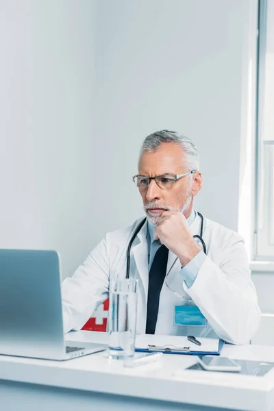 Serious middle aged male doctor using laptop at table in office — Stock Photo