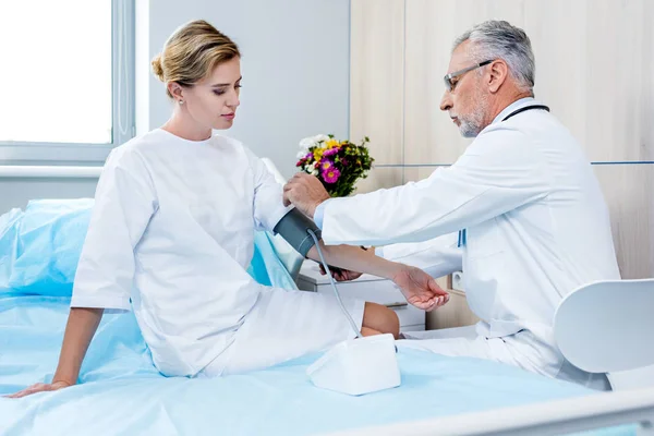 Side view of middle aged male doctor putting on device for measuring pressure on female patient hand in hospital room — Stock Photo