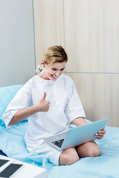 Sonriente paciente femenina haciendo gesto de pulgar hacia arriba y tener videollamada en el portátil en la habitación del hospital - foto de stock