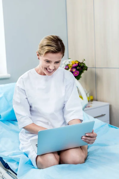 Paciente adulta feliz usando el ordenador portátil en la cama en la habitación del hospital - foto de stock