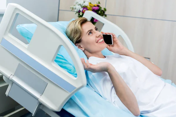 Happy female patient laying on bed and talking on smartphone in hospital room — Stock Photo