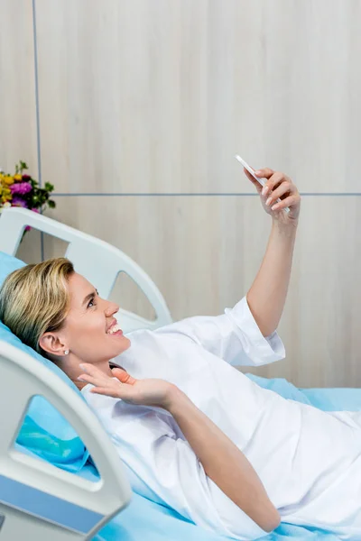 Paciente femenina sonriente saludando a mano y teniendo videollamada en el teléfono inteligente en la habitación del hospital - foto de stock