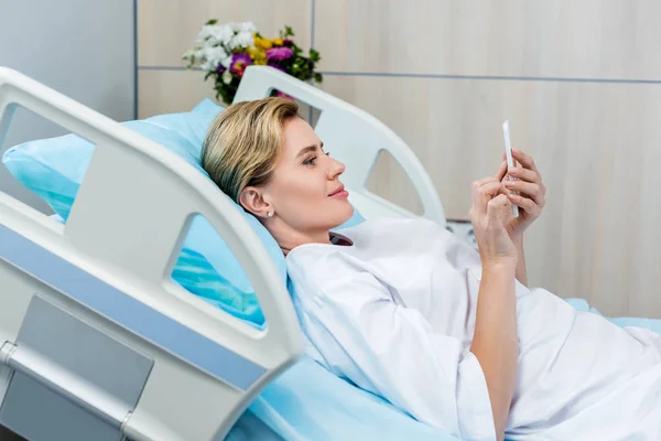 Vista lateral de una paciente adulta feliz usando un teléfono inteligente en la cama en la habitación del hospital - foto de stock