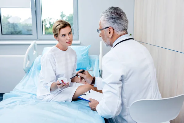 Задний вид врача среднего возраста со стетоскопом над шеей, указывающим на таблетки пациентке в больничной палате — стоковое фото