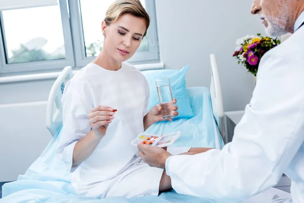 Patiente souriante avec verre d'eau prenant des pilules du médecin masculin mature dans la chambre d'hôpital — Photo de stock