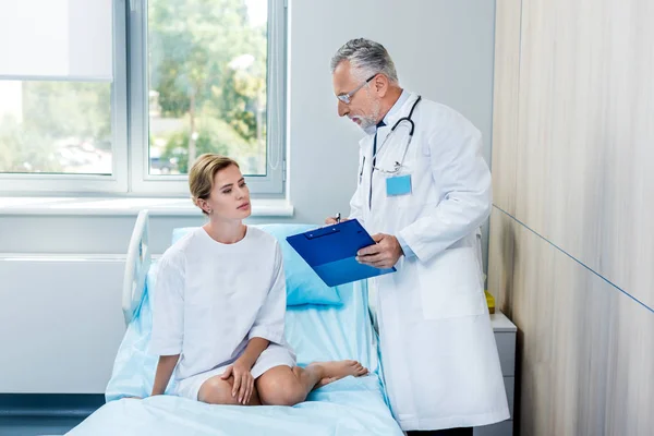 Maturo medico maschio con stetoscopio sopra il collo che punta agli appunti per paziente femminile in camera d'ospedale — Foto stock