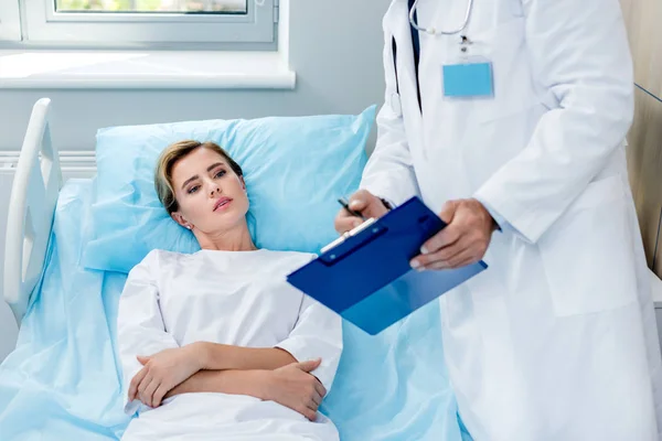 Vue partielle du médecin masculin avec stéthoscope au-dessus du cou pointant vers le presse-papiers du patient féminin dans la chambre d'hôpital — Photo de stock