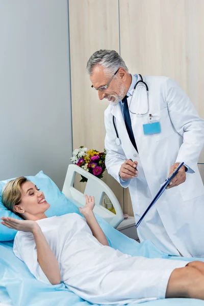 Smiling male doctor with stethoscope over neck holding clipboard and talking to female patient in hospital room — Stock Photo