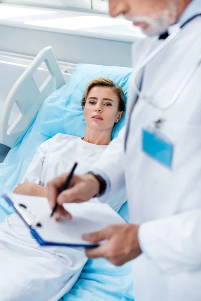 Vista parcial del médico masculino con estetoscopio sobre la escritura del cuello en portapapeles cerca del paciente adulto femenino en la habitación del hospital - foto de stock