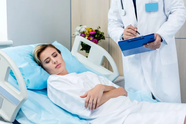 Cropped image of male doctor with stethoscope over neck writing in clipboard near adult female patient in hospital room — Stock Photo