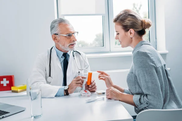 Médico de meia idade do sexo masculino apontando para pílulas para paciente do sexo feminino na mesa no escritório — Fotografia de Stock