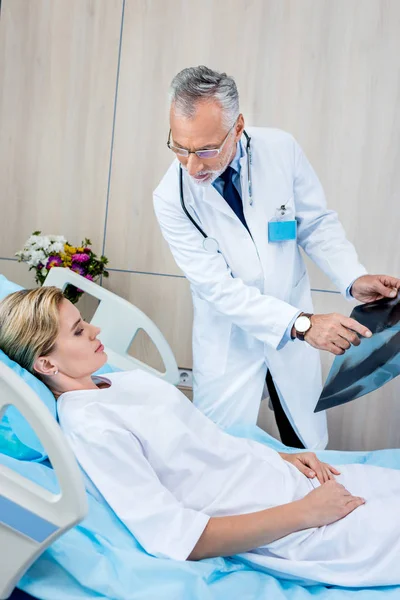 Arzt mittleren Alters zeigt auf Röntgenbild einer Patientin im Krankenhauszimmer — Stockfoto