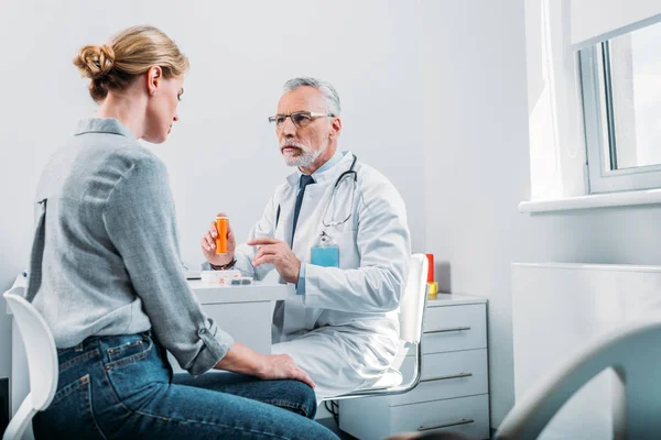 Серьезный зрелый мужчина врач указывая на таблетки к пациентке за столом в офисе — стоковое фото