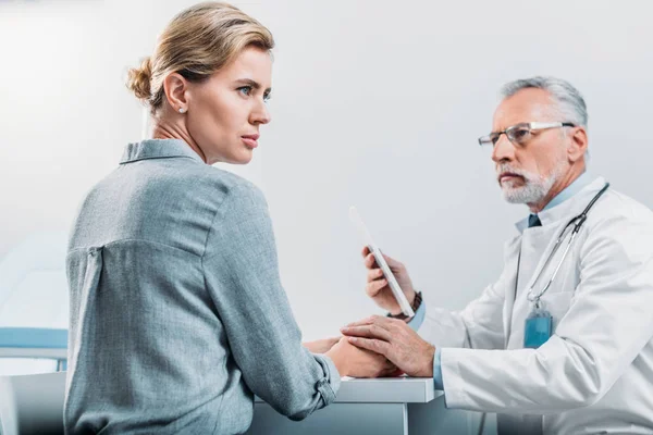 Médico masculino serio con tableta digital animando y toma las manos de paciente mujer molesta en la mesa en el consultorio - foto de stock