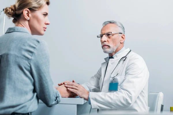Серйозний зрілий чоловік лікар тримає руки пацієнта за столом в офісі — стокове фото