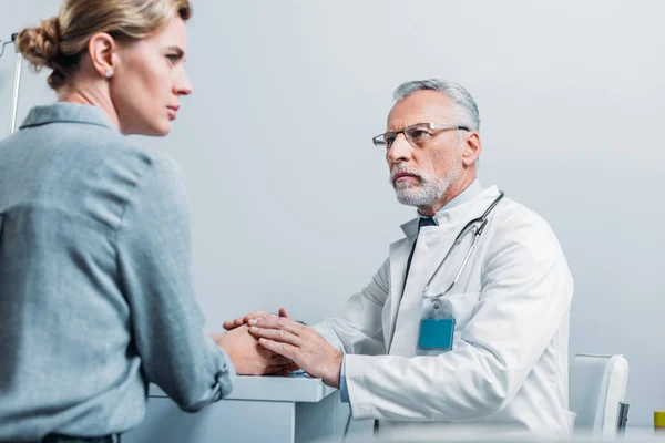 Doctor masculino maduro serio cogido de la mano del paciente femenino en la mesa en el consultorio - foto de stock