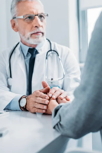 Partial view of middle aged male doctor cheering up and holding hands of female patient at table in office — Stock Photo