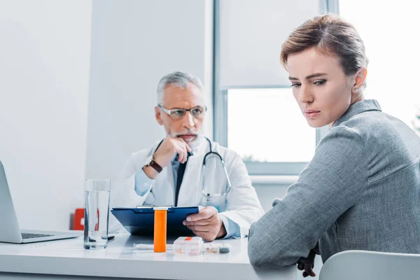 Traurige Patientin schaut weg, während reifer männlicher Arzt in der Nähe am Tisch im Büro sitzt — Stockfoto