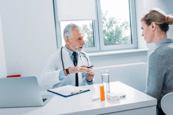 Mature male doctor talking to female patient at table with laptop in office — Stock Photo