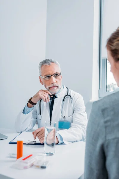 Serious mature male doctor talking to female patient at table with pills and clipboard in office — Stock Photo
