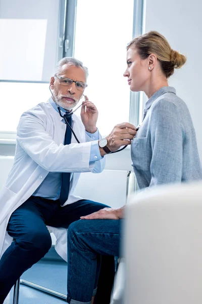 Focused mature male doctor examining female patient by stethoscope in hospital room — Stock Photo