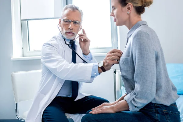 Concentrado maduro masculino médico examinando paciente feminino por estetoscópio no quarto do hospital — Fotografia de Stock