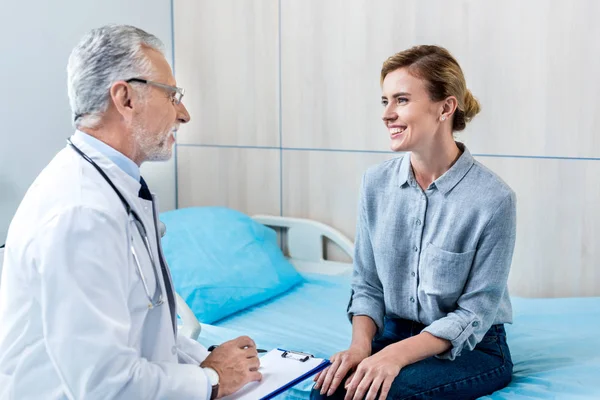 Heureux mature médecin masculin écriture dans le presse-papiers près de patiente dans la chambre d'hôpital — Photo de stock