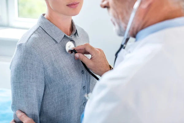 Partial view of mature male doctor examining female patient by stethoscope in hospital room — Stock Photo