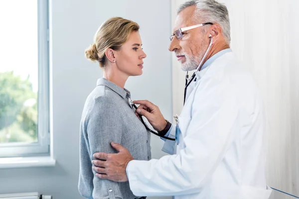 Seitenansicht eines reifen männlichen Arztes, der Patientin im Krankenhauszimmer mit dem Stethoskop untersucht — Stockfoto