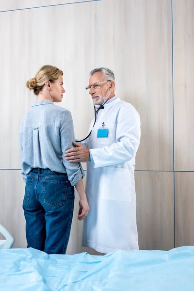 Confident mature male doctor examining female patient by stethoscope in hospital room — Stock Photo