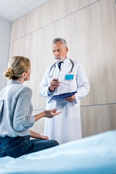 Vista trasera de la paciente femenina gesticulando a mano y hablando con el médico masculino con portapapeles en la habitación del hospital - foto de stock