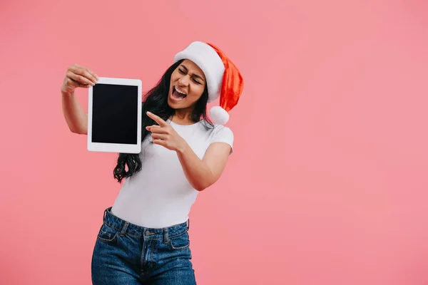 Ritratto di donna afroamericana emotiva in cappello di Babbo Natale che punta al tablet con schermo bianco isolato su rosa — Foto stock