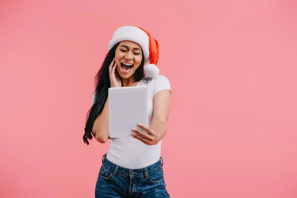 Portrait of emotional african american woman in santa claus hat looking at tablet in hand isolated on pink — Stock Photo