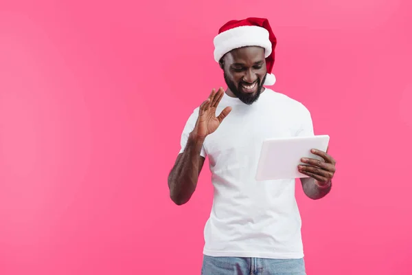 Portrait of smiling african american man in santa claus hat with tablet gesturing isolated on pink — Stock Photo