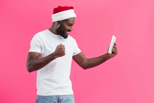Vista lateral del hombre afroamericano sonriente en sombrero de santa claus con tableta aislada en rosa — Stock Photo
