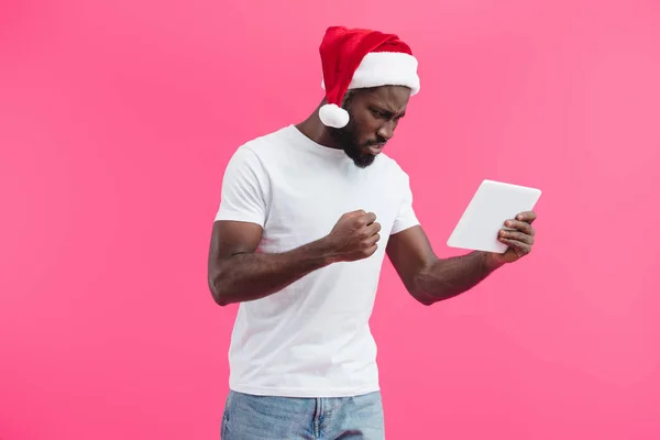 Vue latérale du jeune homme afro-américain en colère dans le chapeau santa claus avec comprimé isolé sur rose — Photo de stock