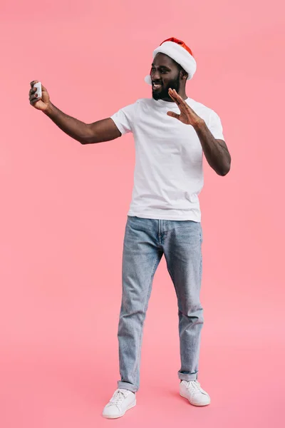 Smiling african american man in santa claus hat with smartphone gesturing on pink background — Stock Photo