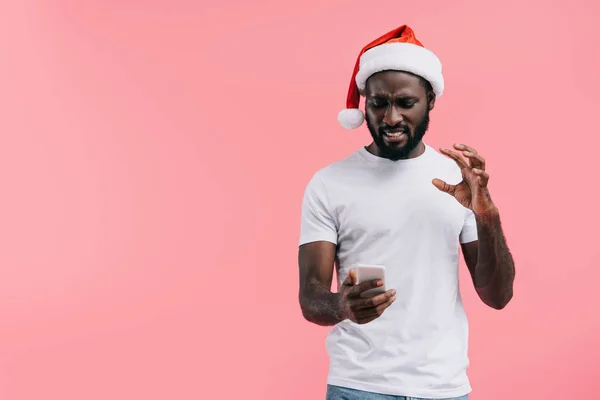 Портрет эмоционального африканского американца в шляпе Санта-Клауса со смартфоном, изолированным на розовом — стоковое фото
