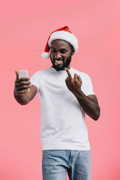 Portrait of smiling african american man in santa claus hat with smartphone showing middle finger isolated on pink — Stock Photo