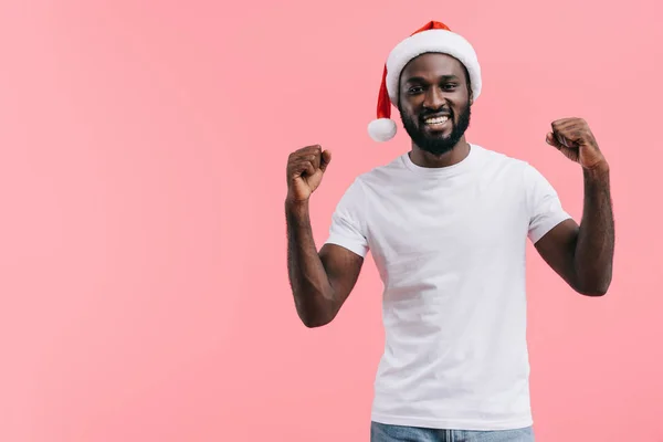 Happy african american man with raised arms in christmas hat isolated on pink background — Stock Photo