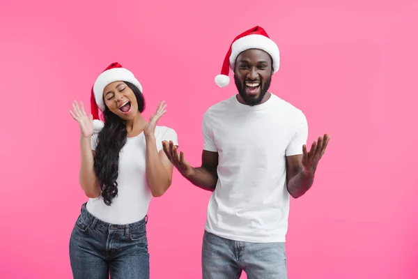 Возбужденная африканская американская пара в рождественских шляпах жестикулируя руками, изолированными на розовом фоне — стоковое фото