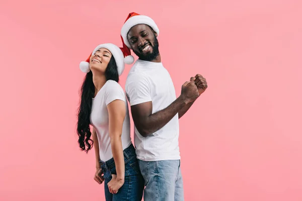 Счастливая африканская американская пара в рождественских шляпах стоя спиной к спине изолированы на розовом фоне — стоковое фото