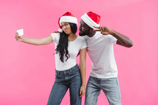 Mujer afroamericana en sombrero de navidad haciendo cara de pato y tomando selfie en smartphone con novio haciendo signo de paz aislado sobre fondo rosa - foto de stock